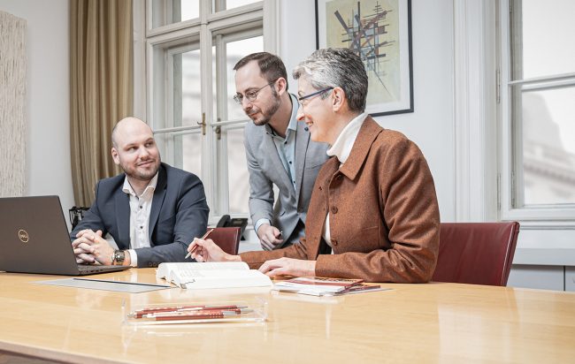 Drei Personen sitzen und stehen um einen Konferenztisch. Eine Frau mit kurzen grauen Haaren in einem braunen Blazer zeigt auf ein offenes Buch. Zwei Männer hören aufmerksam zu. Einer sitzt mit verschränkten Händen vor einem Laptop, der andere lehnt sich ü